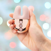 A hand is holding a small, rose-gold Le Wand Chrome Double Motor Vibrator with three buttons on the front against a blurred, colorful background of circles.